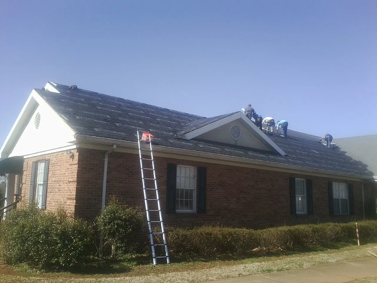 Crew tearing shingles off a brick ranch roof with a ladder set — Metal Roof Repair work by our crew in Minoa