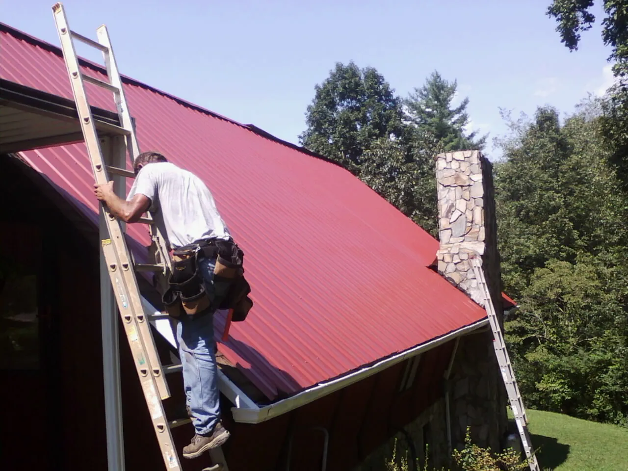 Roofer on a ladder beside a red metal roof and stone chimney — Soffit & Fascia Repair by Roofing Brothers in Minoa